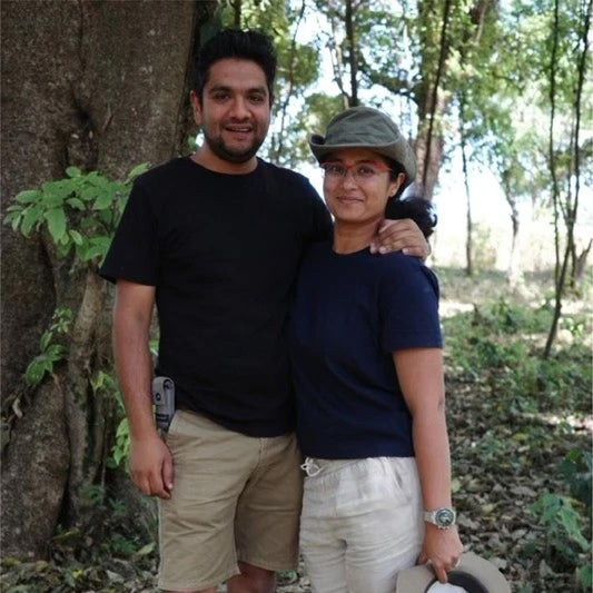 Two people standing together in a forested area with trees and greenery around.