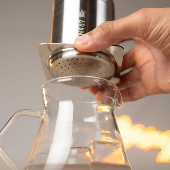 Hand holding a metallic coffee brewing device over a glass container with a blurred background