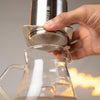 Hand holding a metallic coffee brewing device over a glass container with a blurred background