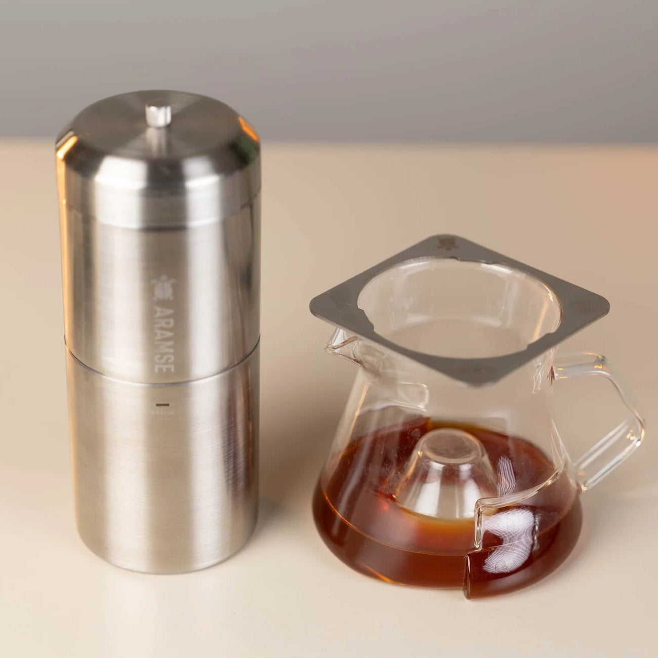 Silver coffee grinder and glass carafe with coffee on a beige surface