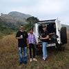 Three people standing in front of a white SUV in a grassy field with mountains in the background.