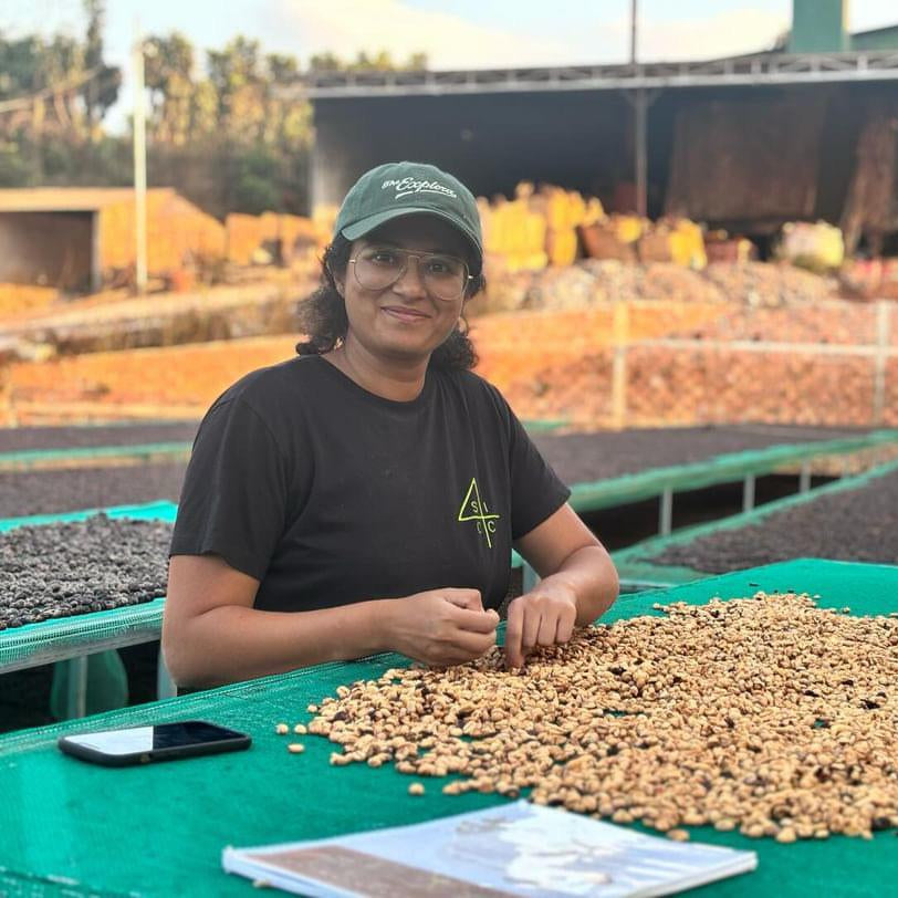 A woman wearing a black t-shirt and green cap sits at a table covered with coffee beans, working in a coffee processing facility with rows of coffee cherries and dark coffee beans in the background.