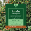 A dark green product label for 'Excelsa Natural process' from Mooleh Manay estate, Coorg, Karnataka, displayed against a background of green and orange coffee plant leaves. The label details the taste profile as tropical fruits, ginger candy, sweet, and smooth, and indicates a roast level of 100% natural.