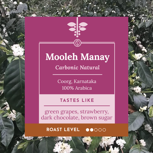 A product label for Mooleh Manay Carbonic Natural coffee, set against a background of green coffee leaves and small white flowers. The label is magenta with white text and a brown bar at the bottom indicating roast level.